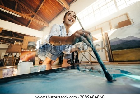 Innovative young artist painting on a large canvas in her studio. Happy female artist looking away cheerfully while working on a new artwork. Smiling woman working on the floor in her atelier. Royalty-Free Stock Photo #2089484860