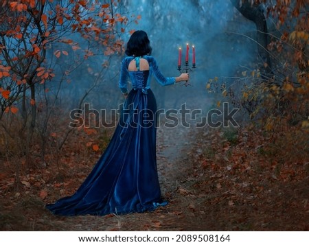 Silhouette fantasy woman queen runs in dark forest, hands hold vintage burning candlestick candles. Blue velvet long medieval dress. Gothic girl vampire princess back rear view. Autumn nature, trees
