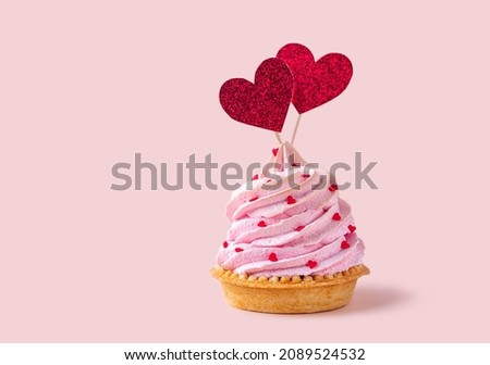 Saint Valentines Day background. Cupcake or tartlet with pink meringue cream decorated with two big red hearts, love symbol, and small heart shape candies. Copy space, pink background.