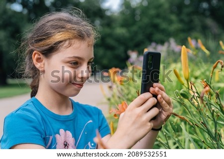 Side view of delighted teenage girl taking picture of blossoming colorful flowers while spending time in park with green trees on summer day