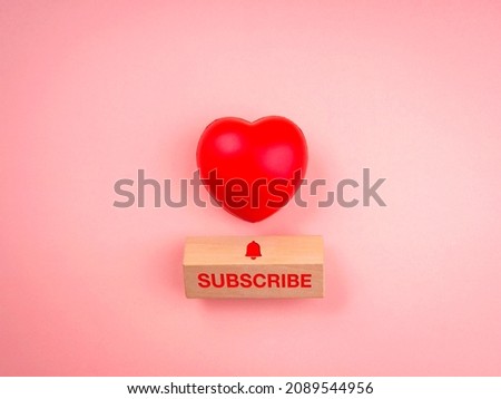 Subscribe concept. Word "SUBSCRIBE" and bell icon on wooden cube block and red heart ball on pink pastel background, minimal style. Following social media.