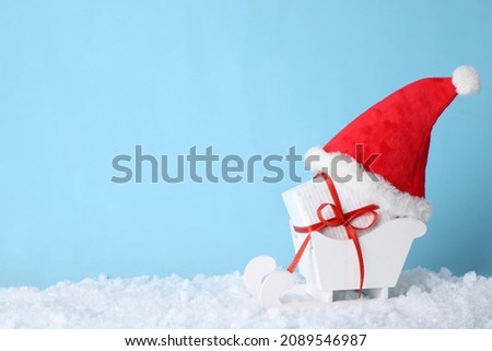 White wooden sleigh with Christmas gift box, Santa hat and artificial snow on light blue background, space for text