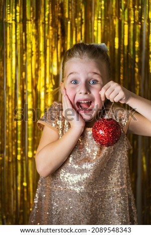 Blonde toddler girl wearing fancy dress hold red Christmas ball against golden glitter background and show her wow emotions