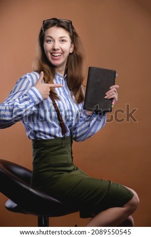 Attractive young Caucasian woman in office style sits on a chair laughing and points to her diary. Studio portrait