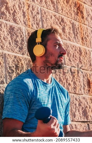 Young adult man training outdoors with dumbbells and yellow wireless headphones.
