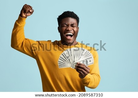 Emotional handsome millennial black man in casual outfit holding bunch of cash, raising hand up and screaming on blue studio background, won lottery, celebrating success, copy space Royalty-Free Stock Photo #2089587310