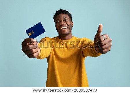 Cheerful young black guy in stylish yellow sweater showing thumb up and credit card, smiling at camera, posing alone on blue studio background, recommending his bank for online shopping or banking Royalty-Free Stock Photo #2089587490