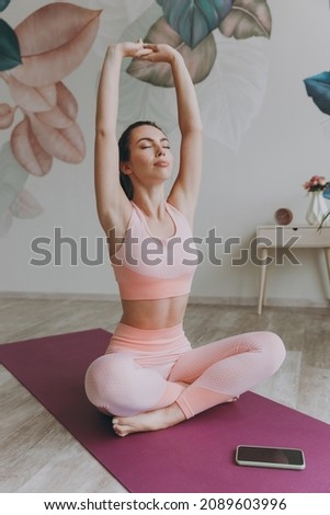 Full body young sporty athletic fitness trainer instructor woman 20s in pink tracksuit doing yoga exercises near mobile cell phone stretch body with hands sit stretch on mat floor at home gym indoor Royalty-Free Stock Photo #2089603996