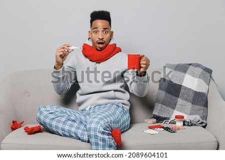 Young shocked african man in grey sweater scarf sit on sofa look at thermometer drink hot tea hold cup isolated on plain gray background studio. Healthy lifestyle ill sick disease treatment concept