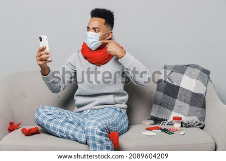 Youngn african man in sweater scarf face mask ppe sit on sofa hold use mobile cell phone talk video call isolated on plain gray background studio. Healthy lifestyle ill sick disease treatment concept