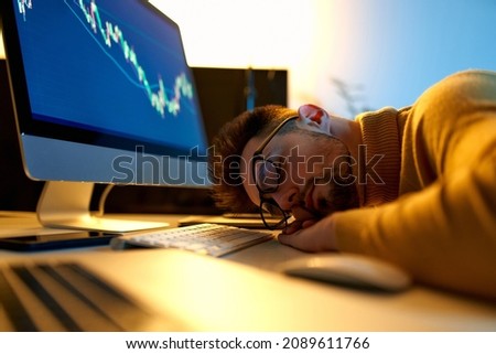 Fatigue man sleeps on his home office desk while trading, browsing online stock investments at night. Tired novice male trader sleeping near monitor with stock chart at workplace. Royalty-Free Stock Photo #2089611766