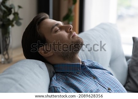 Close up handsome man falls asleep on sofa, put head on soft couch closed eyes breath fresh conditioned air inside modern living room or hotel relaxing indoor. Fatigue relief, climate control concept Royalty-Free Stock Photo #2089628356