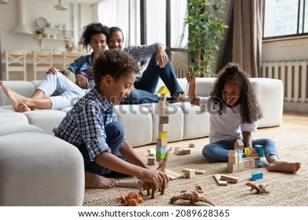 Preschool African siblings play wooden cubes and dino toys sit on warm floor in living room, their parents relaxing on sofa. Family weekend leisure at home, modern playthings, kids development concept Royalty-Free Stock Photo #2089628365