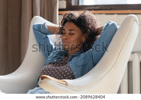Serene African woman put hands behind head sit on leather armchair relaxing inside modern cozy air-conditioned living room. Climate control, fresh air, no stress weekend, fashionable furniture concept Royalty-Free Stock Photo #2089628404