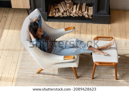 Top view peaceful African woman relax on armchair in modern cottage house with fireplace, put feet on comfy footstool enjoy comfort, rests in warm living room. No stress, air-conditioned home concept Royalty-Free Stock Photo #2089628623
