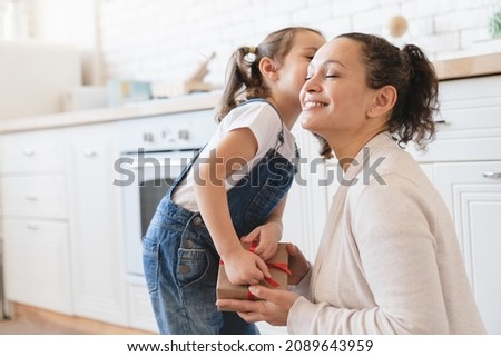 Little cute girl daughter kissing embracing hugging mom giving receiving present gift box for Christmas New Year Mother`s day in the kitchen