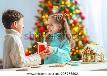 boy gives gift box to little girl. baby is happy and laughs when receiving gift. beautifully decorated room with Christmas tree, gingerbread house on table, boy and girl look at each other and laugh.