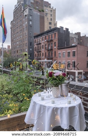A bouquet of flowers sits on top of a table draped with white tablecloth, candles and fairy lights in celebration of a surprise proposal. Buildings in West Villages are in the background.