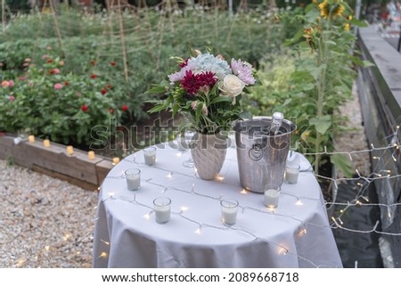 Set up next to a sunflower garden, a bouquet of flowers sits on top of a table draped with a white tablecloth, candles, and fairy lights in celebration of a surprise proposal. 