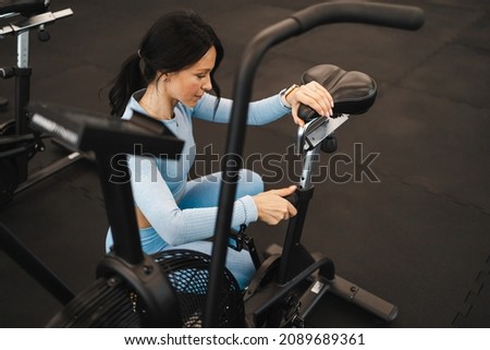 Pretty fitness woman adjusts the height of the exercise bike seat before starting a workout Royalty-Free Stock Photo #2089689361