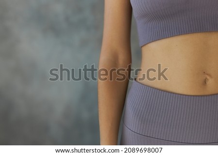 Crop shot of female belly button and hand, dressed in violet short top and leggings, isolated over grey textured wall with copy space for your advertising content. Perfect human body Royalty-Free Stock Photo #2089698007