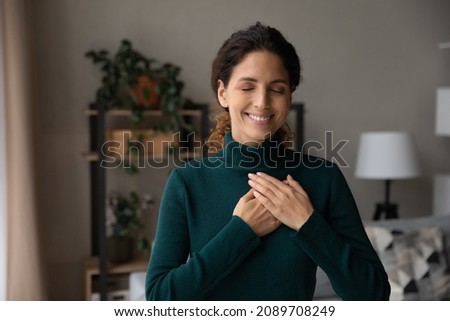 Smiling young Hispanic woman hold hands at heart chest feel grateful and thankful. Happy millennial Latino female believer show love and care support pray or mediate. Faith, superstition concept. Royalty-Free Stock Photo #2089708249