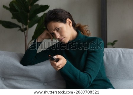 Unhappy millennial Latin woman sit rest on sofa at home feel bored watching TV. Upset sad young Hispanic female renter relax on couch in living room annoyed with television program. Boredom concept.