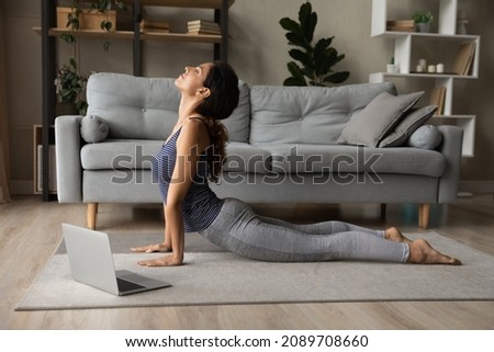 Sporty young Latino woman stretch on floor at home practice yoga or pilates watching video lesson on laptop. Healthy Hispanic female do morning exercise on webcam distant virtual training class.
