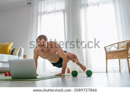 Home workout in the living room. Young muscular body man making spiderman pushups following online a fitness trainer using a laptop. Healthy lifestyle and modern online technology concept.
