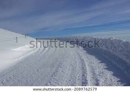 Beautiful winter mountain road covered with snow against the sky with long white clouds. Snowdrifts left on the roadside. Treviso Prealps, Italy.