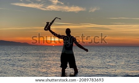 Young male saxophonist stands with his feet in sea water, holds saxophone in his hands, looks at sunset. Beautiful sunset on sea, sky. Musician, playing saxophone, dancing, having fun, Silhouette.