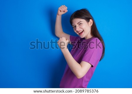 Profile photo of excited Caucasian woman wearing purple T-shirt isolated over blue background raising fists celebrating black Friday shopping