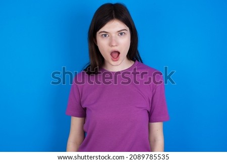 Oh my God. Surprised Caucasian woman wearing purple T-shirt isolated over blue background stares at camera with shocked expression exclaims with unexpectedness,