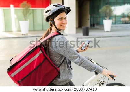 Cheerful woman with a red delivery bag in a bicycle smiling while using her phone to look at the map to make a food delivery 