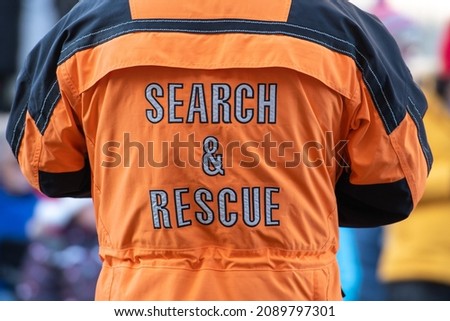 A male adult stands back wearing a bright orange and black safety jacket that has grey lettering. The safety coat has the words search and rescue in the middle of the industrial uniform.