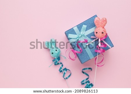 A blue gift box and two Easter bunnies on a stick on a pink background. The concept of Easter holidays. Flat lay.