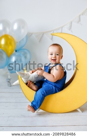 A one-year-old cheerful boy in a blue jumpsuit sits on a yellow wooden crescent next to balloons and a garland of flags.