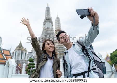 Asian couple backpacker use mobile phone selfie, take picture in city. Attractive man and woman tourist traveler travel on street, using smartphone record vlog on holiday vacation trip in Thailand.