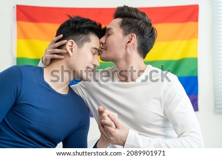 Portrait of Asian handsome man gay family holding LGBT flag and smile. Attractive romantic male lgbt couple feel happy sit on bed in bedroom in morning and kiss with gay pride and rainbow background.