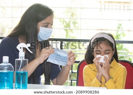 Cautious mother holding medical face mask for her daughter sneezing into tissue at home to protect self from infectious omicron variant of coronavirus disease Royalty-Free Stock Photo #2089908286