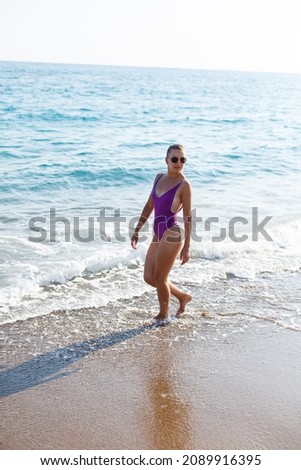 An attractive young woman comes out of the sea in a swimsuit. A girl in a swimsuit walks under the bright summer sun along the sand of the Mediterranean coast. Rest by the sea. Selective focus