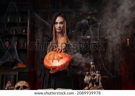 Halloween concept. Black witch holding Halloween pumpkin in hand with carved smily face in hand standing dark room. Female necromancer, dungeon place. Jack o lantern head.