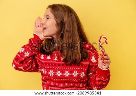 Little caucasian girl holding a Christmas stick isolated on yellow background shouting and holding palm near opened mouth.
