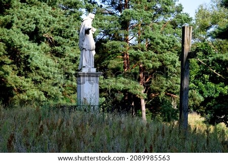 A view of a vast and lush field full of crops with a white marble figurine of a saint standing in the middle of it, next to an old wooden abandoned cross located next to a dense forest or moor