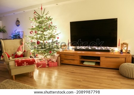 House interior decorated for Christmas with lights on the windows, Christmas tree and a gnome toy in an armchair near the TV in the living room