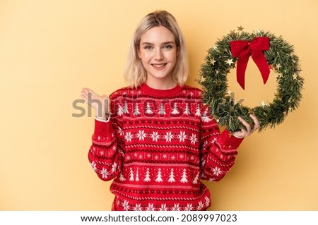 Young caucasian woman holding a Christmas wreath isolated on yellow background showing a copy space on a palm and holding another hand on waist.
