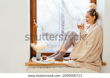 Happy blond woman with bunch hairstyle warming and cover knitted plaid enjoying in her coffee time by the window in cold winter day. Peace of mind and mental health. Royalty-Free Stock Photo #2090000713