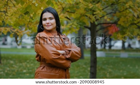 Beautiful young elegant woman looking into distance crossed arms over chest turns slowly focuses gaze on camera smiles shyly posing attractive pretty Hispanic girl stand in city autumn park outdoors