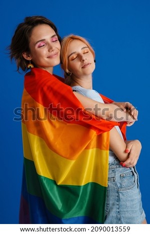 White lesbian couple hugging while posing with rainbow flag isolated over blue background