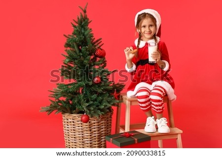 Little girl in Christmas costume with cookie and glass of milk on red background